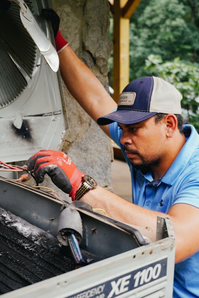 A man wearing gloves and a cap repairs an open outdoor air conditioning unit, inspecting wires and components with a focused expression.