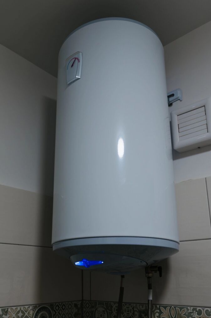 A white cylindrical water heater is mounted high on a bathroom wall, with indicator lights illuminated and pipes connected at the bottom.