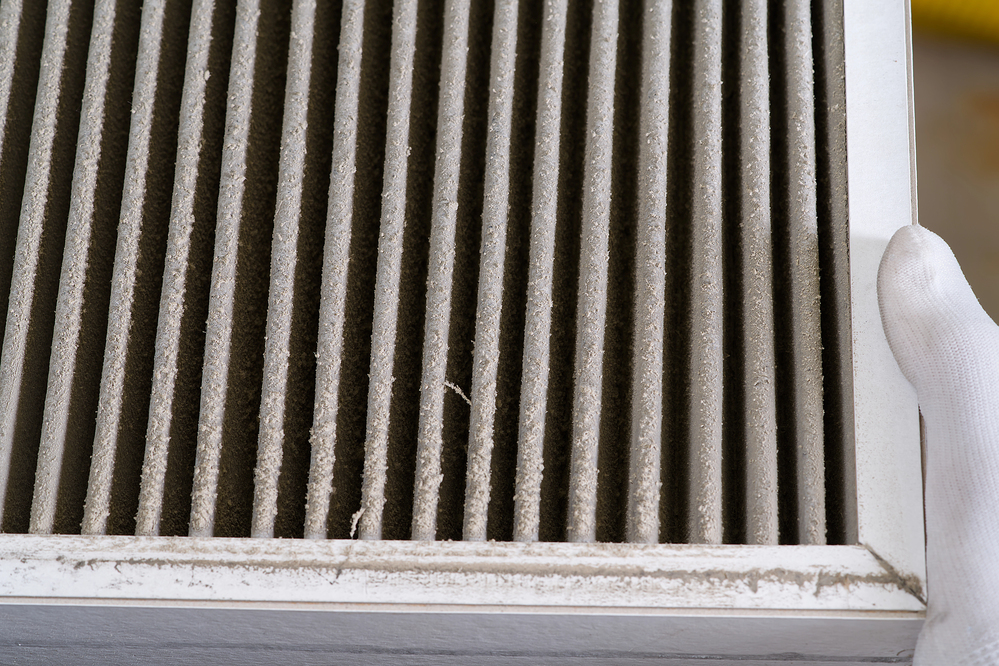 A close-up of a dirty air filter with visible dust buildup, held by a person wearing a white glove.