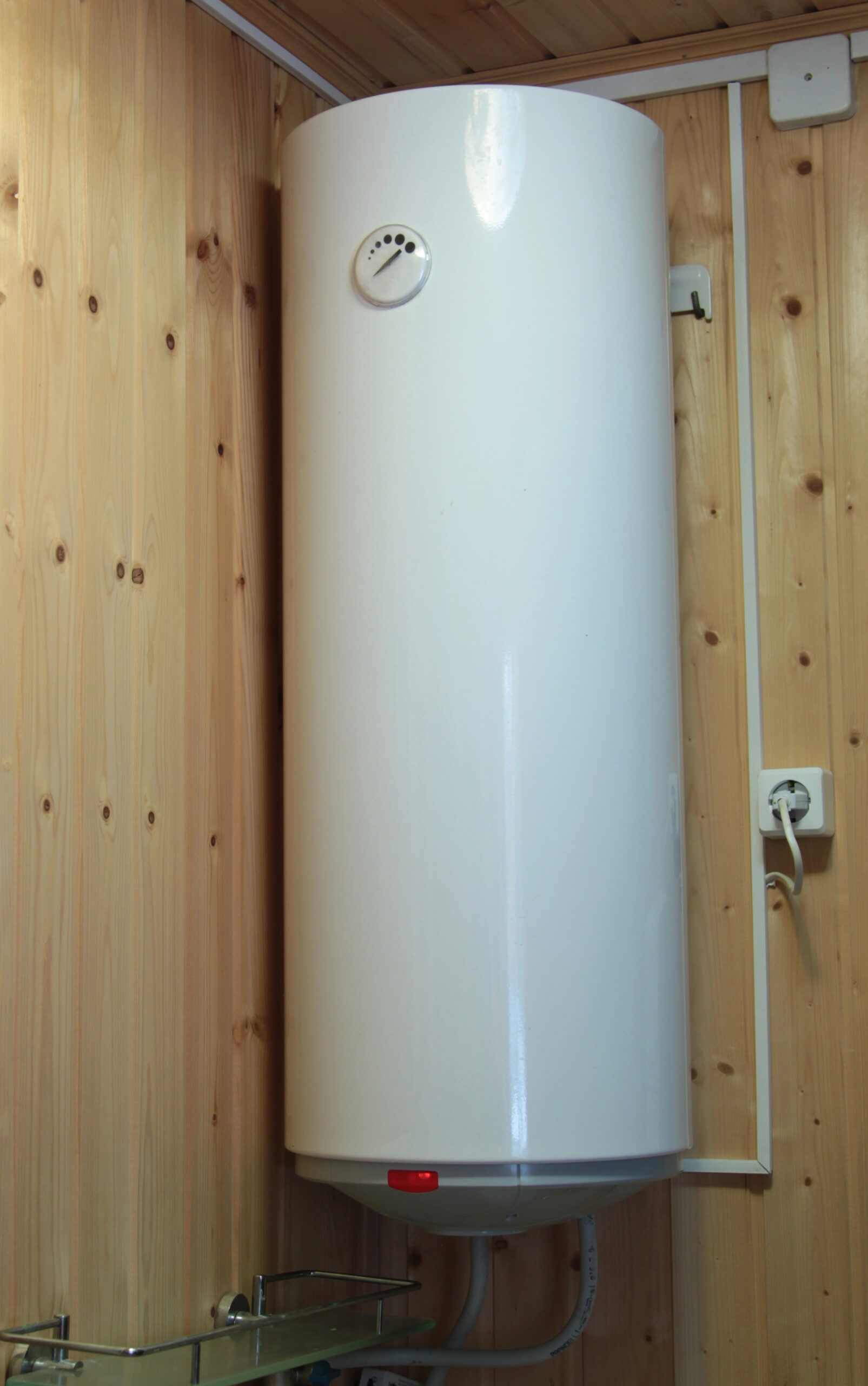 A white cylindrical water heater mounted vertically on a wooden wall, with visible plumbing and electrical connections.